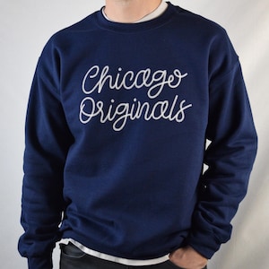 Puede incluir: Sudadera azul marino con la inscripci&oacute;n "Chicago Originals" en blanco en el pecho. La sudadera tiene cuello redondo y mangas largas. La persona lleva una camiseta interior blanca.