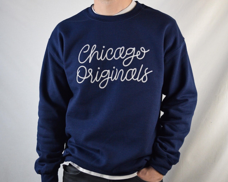 Puede incluir: Sudadera azul marino con la inscripci&oacute;n "Chicago Originals" en blanco en el pecho. La sudadera tiene cuello redondo y mangas largas. La persona lleva una camiseta interior blanca.