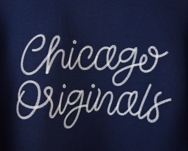 Puede incluir: Tela azul marino con las palabras "Chicago Originals" bordadas en blanco con escritura cursiva. El texto est&aacute; en una fuente decorativa y fluida, creando un dise&ntilde;o elegante.