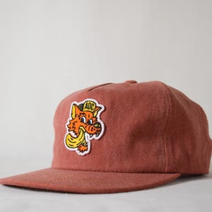 May include: A red baseball cap with a patch of a cartoon fox wearing a hat that says "ADC" on it.