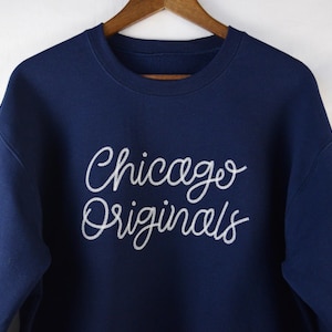Puede incluir: Sudadera azul marino con la inscripci&oacute;n "Chicago Originals" en escritura blanca en el pecho. La sudadera est&aacute; colgada de una percha de madera. La parte superior de manga larga es de material suave.