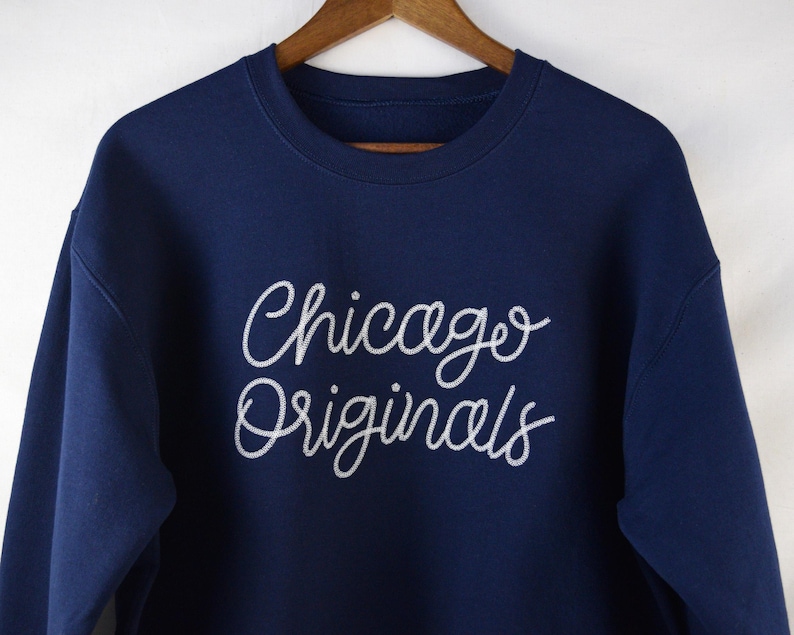 Puede incluir: Sudadera azul marino con la inscripci&oacute;n "Chicago Originals" en escritura blanca en el pecho. La sudadera est&aacute; colgada de una percha de madera. La parte superior de manga larga es de material suave.