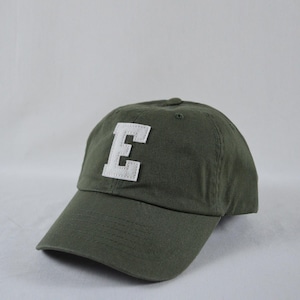 May include: An olive green baseball cap featuring a large, white, block-letter E patch centered on the front panel. The cap has a curved brim, stitched detailing, and a structured crown with ventilation eyelets on the top panels.
