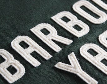 May include: A dark forest green fabric featuring the word BARBOUR embroidered in raised, off-white, corded lettering. The stitching creates a textured, three-dimensional effect across the surface of the dark green textile material.