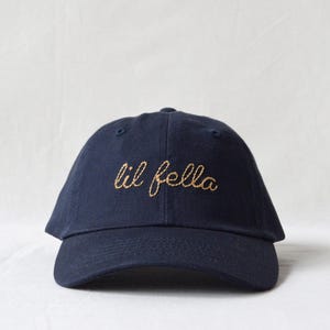 May include: A navy blue baseball cap featuring the words lil fella embroidered in a gold-colored cursive script across the front panels. The cap has a curved brim, a structured crown with eyelets, and a button at the top center of the headwear.