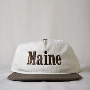 May include: A white baseball cap with a brown brim. The word "Maine" is embroidered in brown block letters across the front of the cap. The cap is made of a canvas-like material.