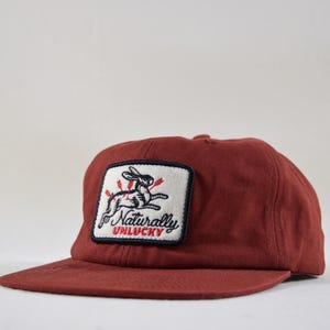 May include: A maroon baseball cap with a flat brim. The cap features a rectangular patch with a rabbit pierced by arrows, and the words "Naturally Unlucky" in a stylized font. The cap is made of a durable fabric.