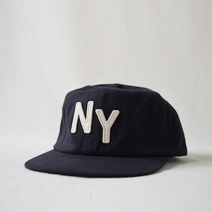 May include: A dark navy blue baseball cap featuring the letters Ny in white, textured, stitched-on fabric on the front panel. The cap has a structured crown and a flat brim, constructed from a solid-colored fabric with visible stitching details.