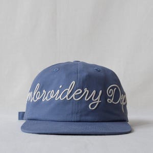 May include: A dusty blue baseball cap featuring the white embroidered text "Embroidery Dep" in a cursive script across the front panels. The cap has a structured crown, a flat brim, and a fabric strap closure visible on the left side.