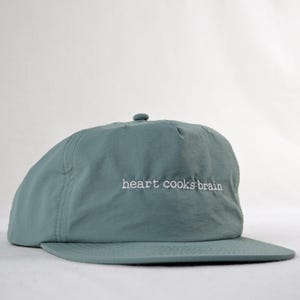 May include: A teal baseball cap with a curved peak. The phrase "heart cooks brain" is embroidered in white on the front. The cap is made of a smooth, slightly shiny fabric.