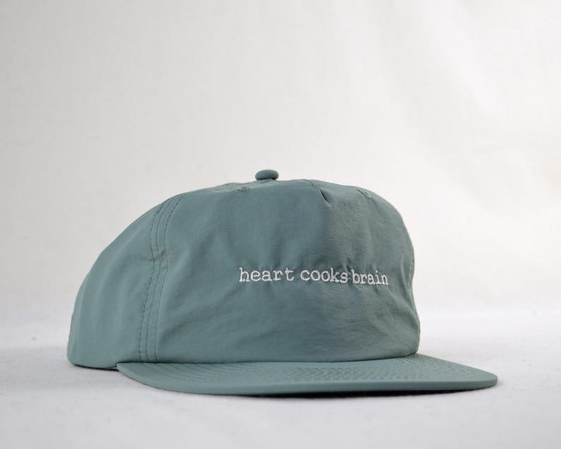 May include: A teal baseball cap with a curved peak. The phrase "heart cooks brain" is embroidered in white on the front. The cap is made of a smooth, slightly shiny fabric.