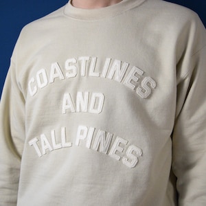 May include: A beige sweatshirt with the text "COASTLINES AND TALL PINES" in white raised lettering.