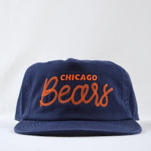 May include: Navy blue baseball cap with the words "CHICAGO Bears" embroidered in orange. The cap has a flat brim and a classic, adjustable fit. A stylish accessory for sports fans.