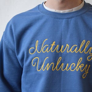 May include: A blue crewneck sweatshirt featuring the text Naturally Unlucky embroidered in yellow cursive script across the chest. The garment is worn over a white collared shirt, showcasing a classic long-sleeve design with ribbed cuffs.