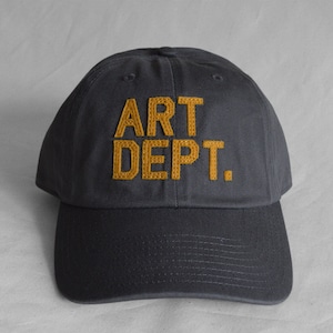 May include: A dark gray baseball cap with the words "ART DEPT." embroidered in yellow on the front.