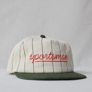 May include: An off-white baseball cap featuring thin vertical dark green pinstripes, a solid dark green brim, and a dark green button on top. The word sportsman is embroidered in red cursive script across the front center of the cap.