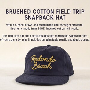 Puede incluir: Gorra snapback azul marino con bordado dorado "Redondo Beach". Cuenta con una visera curva y cierre de broche de pl&aacute;stico ajustable. El texto "BRUSHED COTTON FIELD TRIP SNAPBACK HAT" est&aacute; en la parte superior.