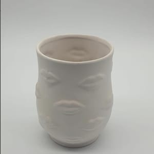 Może przedstawiać: Biały ceramiczny wazon o cylindrycznym kształcie. Wazon zdobiony jest powtarzającym się wzorem wypukłych kształtów ust. Wnętrze wazonu jest w jaśniejszym odcieniu bieli. Ten dekoracyjny wazon nadaje się do dekoracji wnętrz.