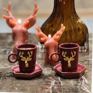 May include: Two burgundy ceramic coffee cups with gold deer accents and matching saucers. The cups are filled with coffee and are sitting on a dark brown surface.