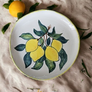 May include: A white ceramic plate with a yellow and green lemon design. The plate has a yellow rim and is decorated with lemons and leaves.