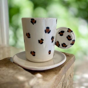 Op de afbeelding: Een witte keramische koffiekop met een bruin en zwart luipaardprintontwerp. De kop heeft een unieke, afgeronde handgreep die ook is versierd met de luipaardprint. De kop staat op een witte schotel.