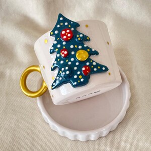 May include: A white ceramic mug with a blue and green Christmas tree design. The tree is decorated with red and yellow ornaments and white dots. The mug has a gold handle and comes with a matching saucer.