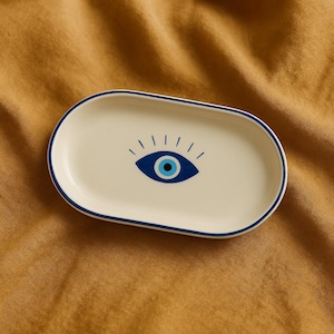 Puede incluir: Plato ovalado de cerámica blanca con un diseño de ojo azul malvado. El plato tiene un borde azul.