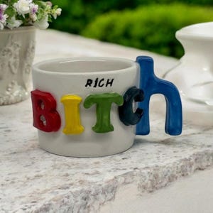 May include: White ceramic mug with the word "BITCH" spelled out in colourful, raised letters. The handle is shaped like the letter "H" and is blue. The mug also has the word "RICH" written on the side.