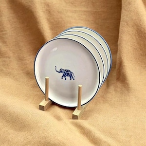 May include: Set of four white ceramic plates with a blue geometric elephant design and a blue rim. The plates are stacked on a wooden stand.