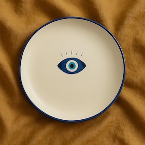 May include: A white ceramic plate with a blue evil eye design. The plate has a dark blue rim.