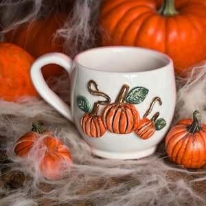 May include: White ceramic mug with a autumnal theme. The mug features a raised design of three pumpkins with green leaves and stems. The pumpkins are orange and brown.