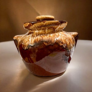 May include: A ceramic lidded jar featuring a glossy finish with a deep brown base and a cream-colored drip glaze pattern cascading from the rim. The vessel has a wide opening, a matching lid with a central knob, and two small side handles.