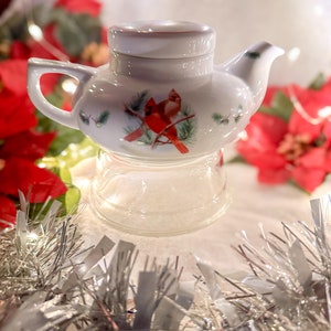 Peut inclure: Une théière en céramique blanche avec couvercle, ornée d'une illustration décorative de deux cardinaux rouges perchés sur des branches de pin. La théière est posée sur une base en verre transparent, avec une finition lisse et brillante.