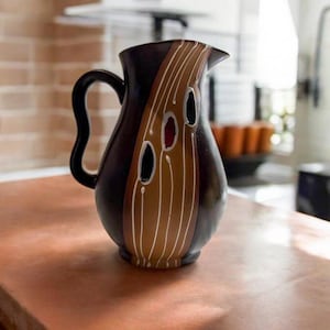 Könnte beinhalten: Eine braune Keramikkanne mit einem goldenen und weißen abstrakten Design. Die Kanne hat einen geschwungenen Griff und eine breite Öffnung.