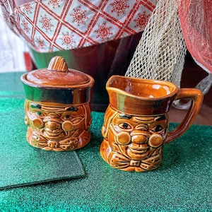 Puede incluir: Un juego vintage de jarra de crema y azucarero de cerámica. El juego presenta un diseño de cara sonriente con un esmalte marrón, detalles verdes y una tapa en el azucarero. La jarra de crema tiene un asa y un pico. El juego está sobre una superficie verde.