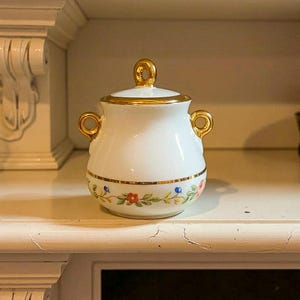 Peut inclure: Un sucrier en porcelaine blanche avec une poignée et une bordure dorées. Le sucrier présente un motif floral avec des fleurs rouges, bleues et roses. Il a deux poignées dorées sur les côtés.