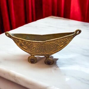 May include: An antique brass-coloured bowl with a boat-like design. The bowl has intricate floral and circular patterns, and small wheels at the base. The handles are curved, and it sits on a white marble surface.