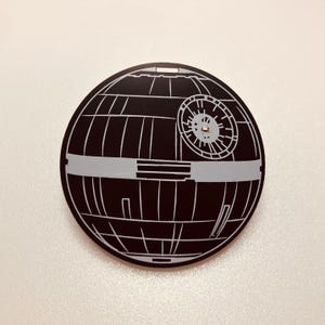 May include: A circular black and light gray flat ornament featuring a detailed graphic design of a spherical space station. The design includes a prominent circular dish on the upper right side, a horizontal band across the center, and vertical lines.
