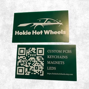May include: Two rectangular dark green cards with white text and graphics. The top card features a stylized car outline and the text Hokie Hot Wheels. The bottom card displays a QR code, the text CUSTOM PCBS, KEYCHAINS, MAGNETS, LEDS, and the URL https://hokiehotwheels.etsy.com.
