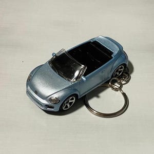 Puede incluir: Un llavero de coche descapotable azul claro con un llavero plateado. El coche de juguete tiene un diseño detallado, que incluye faros, ruedas y un parabrisas. El coche es una réplica en miniatura de un Volkswagen Beetle.
