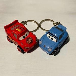 Puede incluir: Dos llaveros de coches de juguete. Uno es rojo con el número 95 y la palabra "Rusteze". El otro es un coche azul. Ambos tienen llaveros plateados.