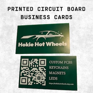 May include: Two rectangular green printed circuit board business cards. The top card features a white car graphic and the text Hokie Hot Wheels. The bottom card displays a white QR code and the text CUSTOM PCBS, KEYCHAINS, MAGNETS, LEDS, and the URL.