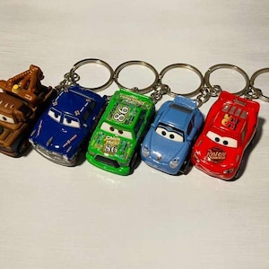 May include: A set of five character keychains featuring miniature toy cars. The collection includes a brown tow truck, a blue car with number 51, a green car with number 86 and text Chick Hicks, a light blue car, and a red car with number 95 and Rust-eze text.