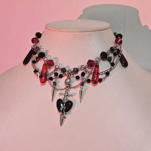 May include: A black and silver choker necklace with red glass beads, a skull charm, and a dagger charm with a black heart.