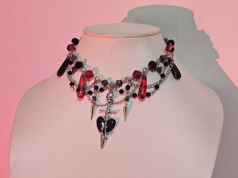 May include: A black and silver choker necklace with red glass beads, a skull charm, and a dagger charm with a black heart.