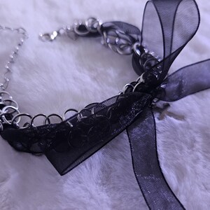 May include: A silver chain choker necklace with a black ribbon bow. The ribbon is made of sheer fabric and has a sparkly finish.