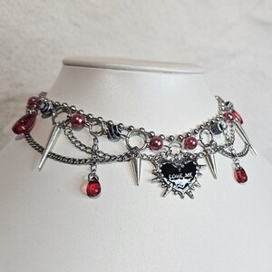 May include: A silver choker necklace with multiple chains, red and black beads, and silver spikes. A black heart charm with the words "LOVE ME" is hanging from the center of the necklace.