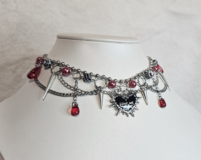 May include: A silver choker necklace with multiple chains, red and black beads, and silver spikes. A black heart charm with the words "LOVE ME" is hanging from the center of the necklace.