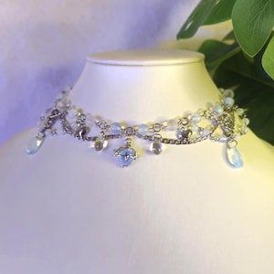 May include: A silver chain choker necklace with light blue and white beads and charms. The necklace has a delicate and feminine design.