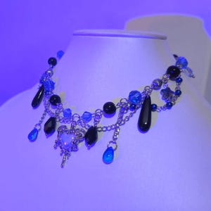 May include: A silver chain necklace with black and blue beads, a silver charm, and dangling blue and black teardrop-shaped beads.
